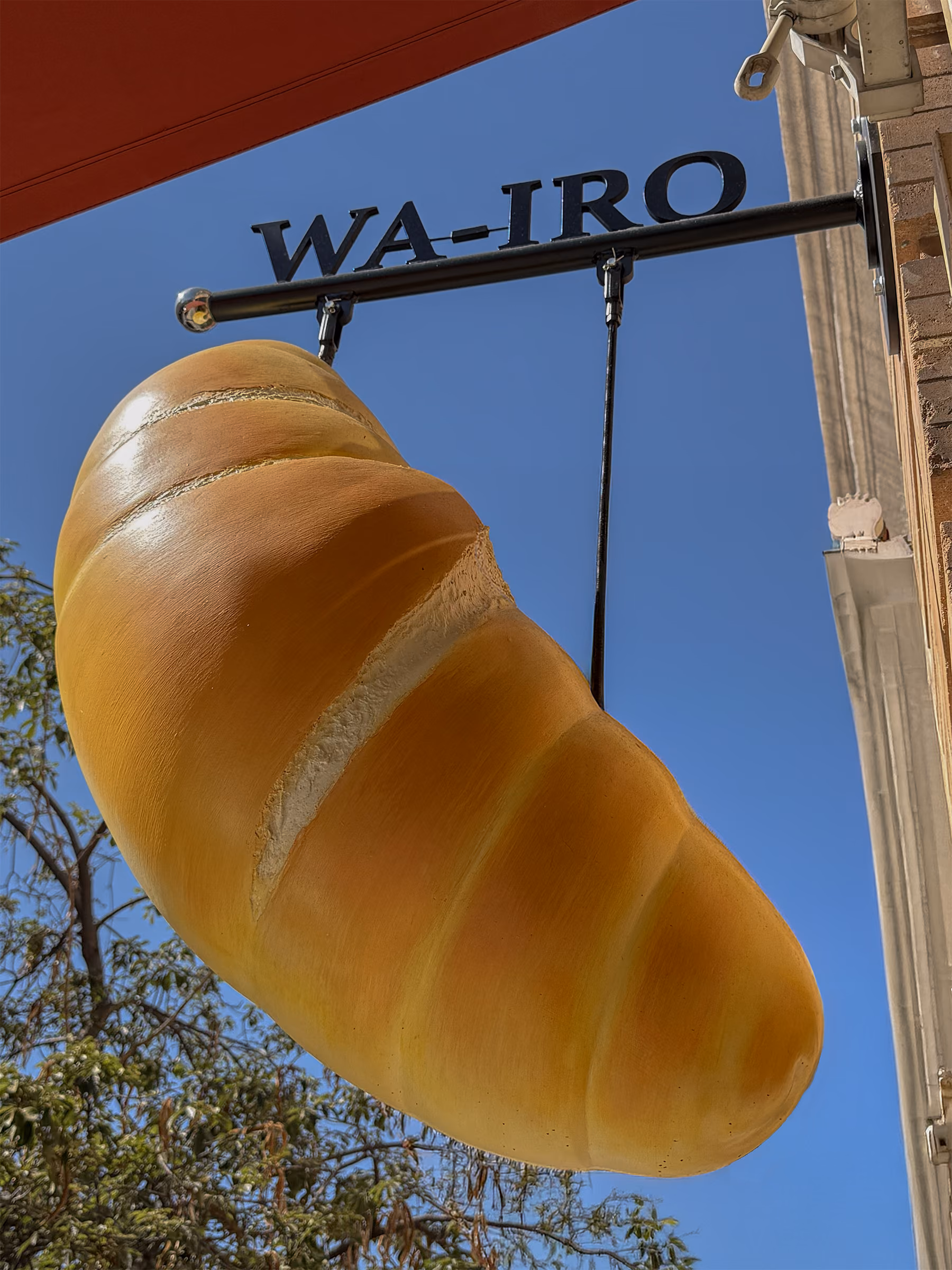 A large sculptural croissant hanging beneath the WA-IRO blade sign on the storefront, against a clear blue sky.