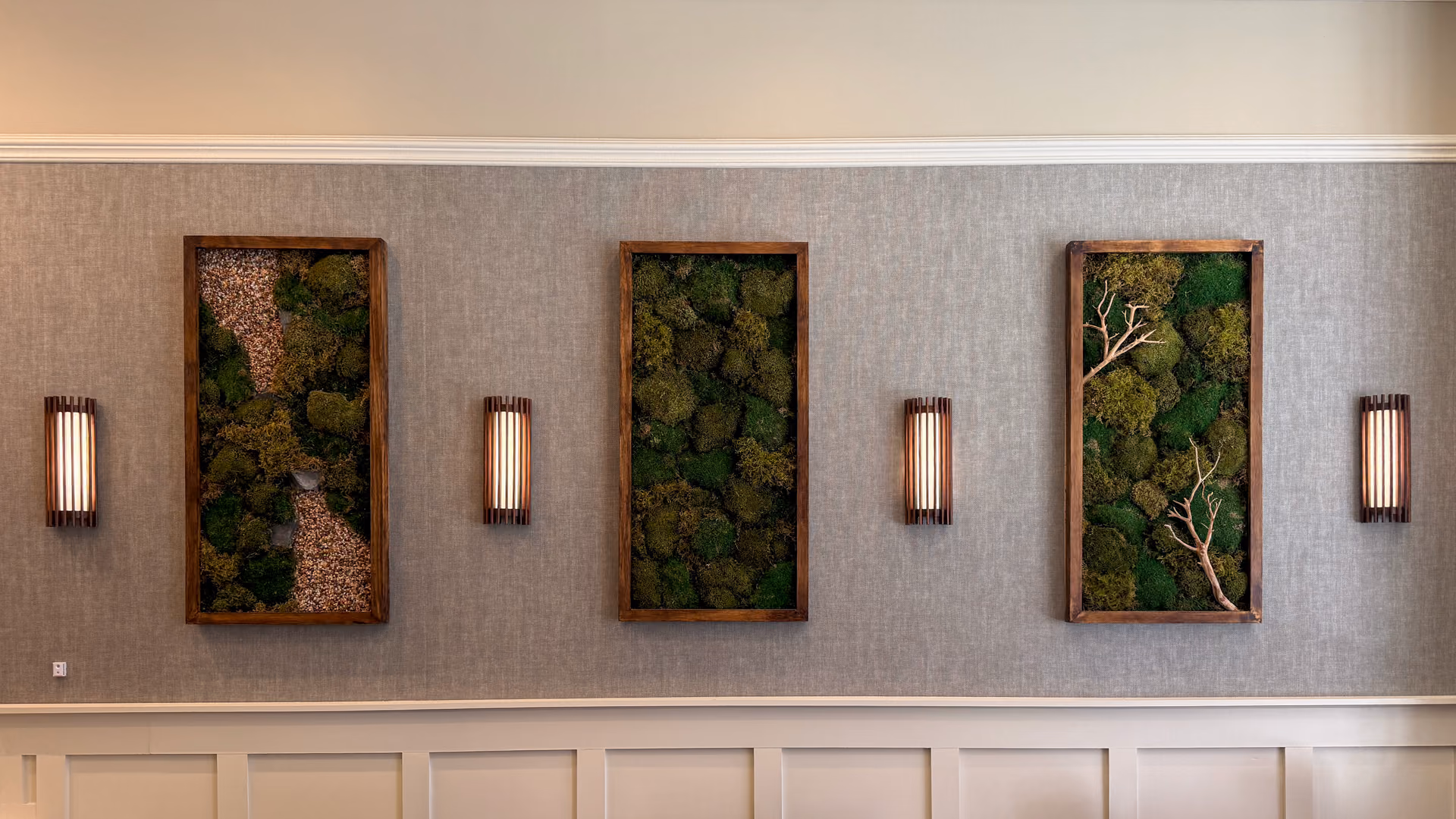 Three framed panels of preserved moss, arranged as miniature Japanese garden scenes with gravel and bare branches, hanging between slatted lantern-style wall sconces inside the café.
