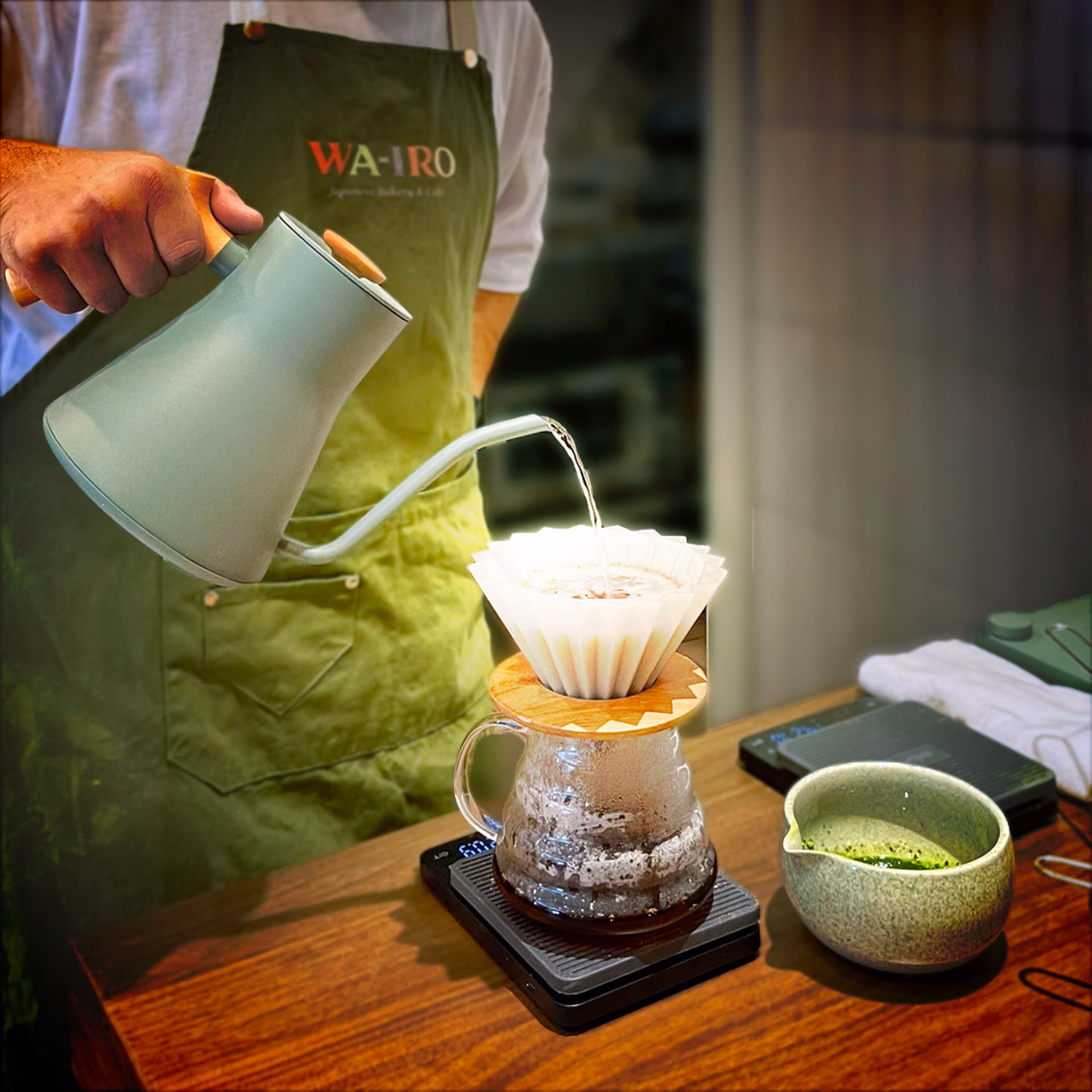 A staff member pouring hot water over a paper filter into a glass carafe, a bowl of whisked matcha beside it.