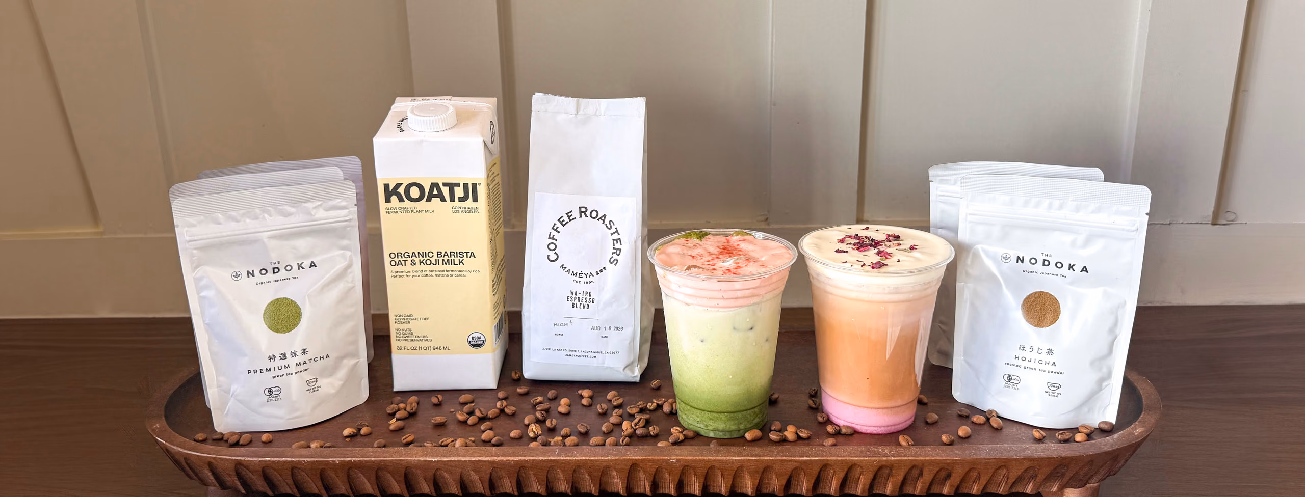 A styled lineup on a wooden tray — Nodoka premium matcha and hojicha pouches, a carton of Koatji organic barista oat and koji milk, a Mameya Coffee Roasters espresso blend bag, and two iced drinks, with coffee beans scattered across the tray.