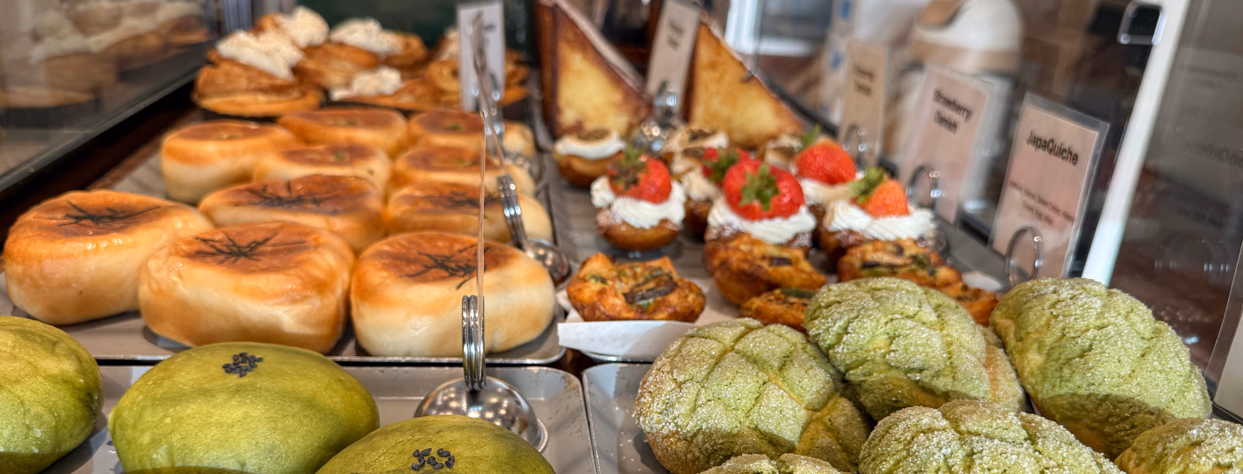 A bakery case seen down its length — glossy cream buns, matcha melon pan, strawberry-topped danishes, small savory quiches and thick-cut French toast filling tray after tray.
