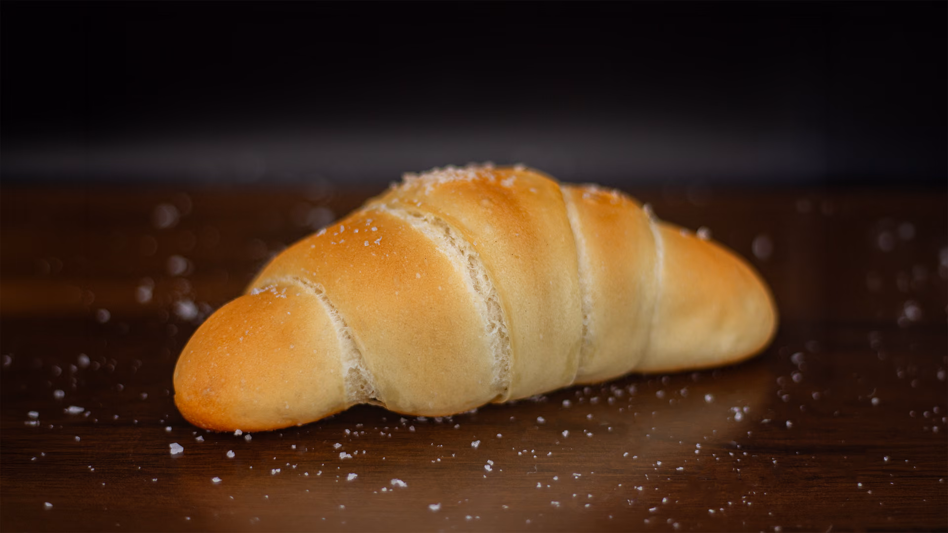 A golden salt-and-butter roll on dark walnut, flaked salt scattered around it.