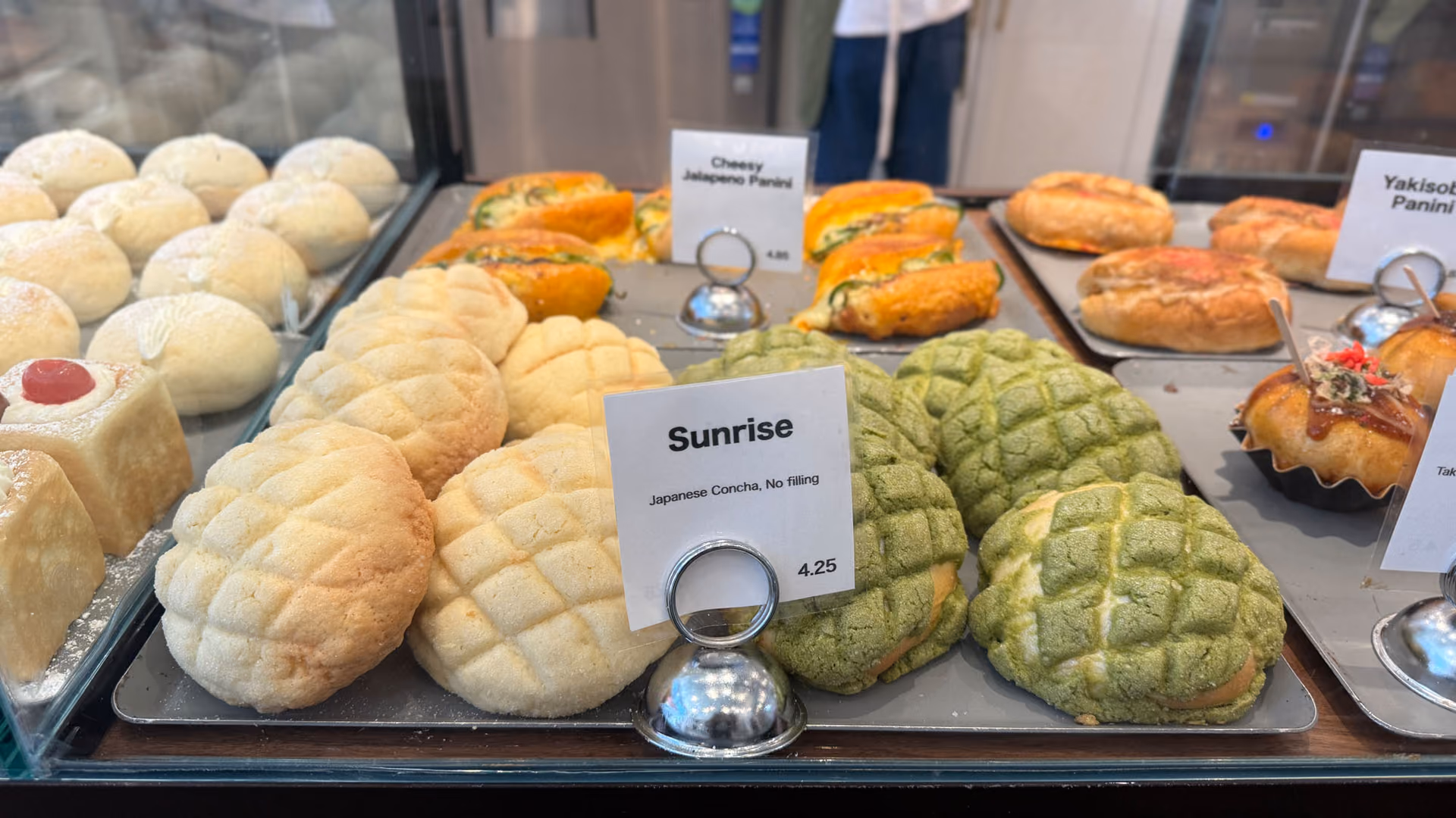 Plain and matcha Japanese melon pan (concha) in the pastry case, a "Sunrise" price tag reading "Japanese Concha, No filling, $4.25."